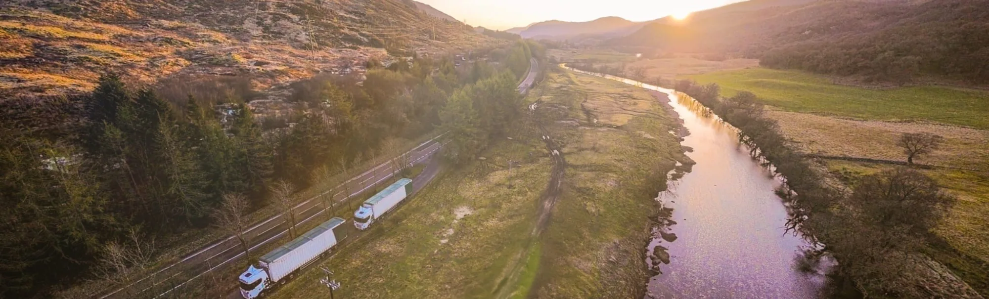 Drone angle scene of the river with the sun setting in the distance and two A.W. Jenkinson wagons parked in the lay-by next to the road