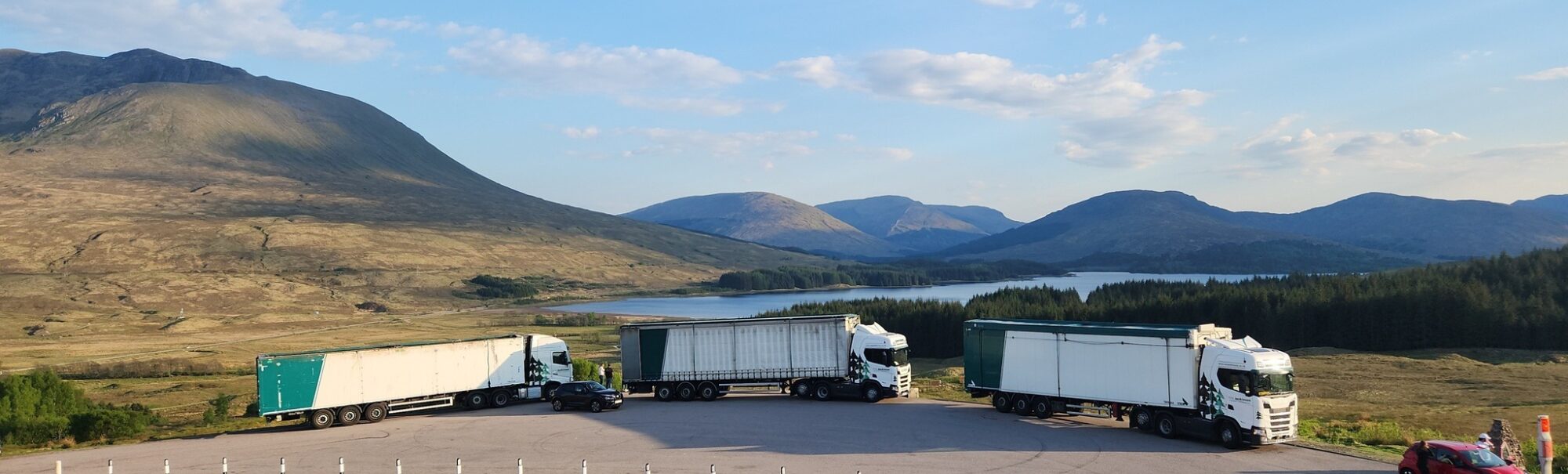 Three white and green A.W. Jenkinson wagons parked in front of a scenic loch in the Scottish highlands.
